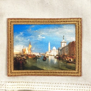 May include: A framed embroidered patch depicting a cityscape with a canal, boats, and buildings. The sky is blue with white clouds. The frame is gold with a brown border.