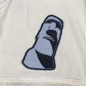 May include: A gray and black embroidered patch of the Moai statue from Easter Island.