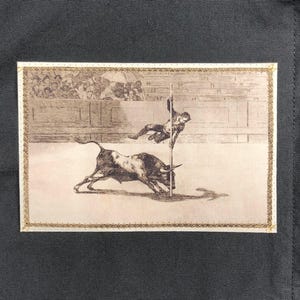 May include: A sepia-toned print depicting a bullfight scene. A bull charges towards a figure on a pole, with a crowd in the background. The print is framed with a gold border and set against a dark gray fabric.