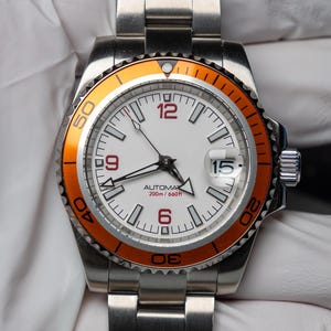 Puede incluir: Reloj de pulsera de acero inoxidable con esfera blanca y bisel naranja. Tiene una pantalla de fecha y una correa de acero inoxidable. La esfera muestra el texto "AUTOMATIC 200m/660ft".