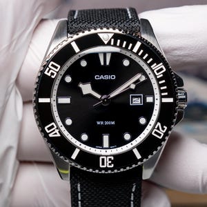 May include: A black Casio wristwatch with a stainless steel case, a black rotating bezel with white numerals, and a black dial featuring luminous markers and hands. The watch includes a date window at the three o'clock position and a black fabric strap.