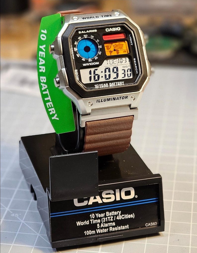 May include: A digital Casio watch with a silver case, brown leather strap, and a black display showing the time. The watch is on a black display stand with the Casio logo and text. A green tag reads "10 YEAR BATTERY".