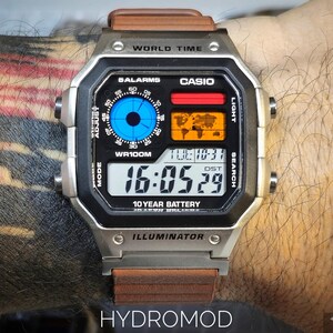 May include: A silver digital watch with a brown leather strap. The watch face displays the time, date, and world map. The watch has the text "WORLD TIME", "CASIO", "10 YEAR BATTERY", and "HYDROMOD".