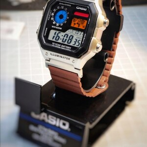 May include: A Casio digital watch with a brown leather strap is displayed on a black stand. The watch face shows the time, date, and various functions. The watch has a silver case and black face with the word "ILLUMINATOR" at the bottom.