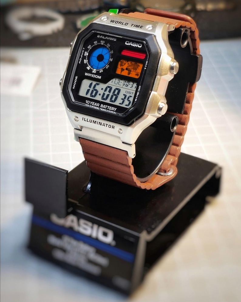 May include: A Casio digital watch with a brown leather strap is displayed on a black stand. The watch face shows the time, date, and various functions. The watch has a silver case and black face with the word "ILLUMINATOR" at the bottom.