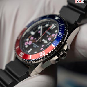 May include: Close-up of a black wristwatch with a black band. The watch face is black with pink floral accents and white hands. The bezel is red and blue. The watch has a stainless steel case and a black rubber strap. The text "Watches World" is in the top right corner.