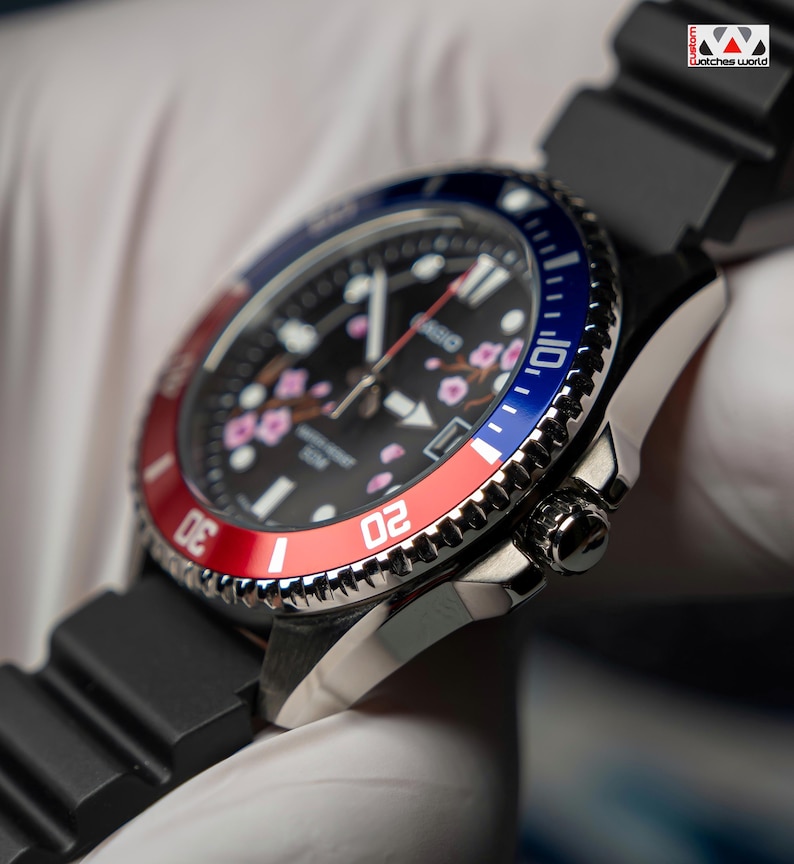 May include: Close-up of a black wristwatch with a black band. The watch face is black with pink floral accents and white hands. The bezel is red and blue. The watch has a stainless steel case and a black rubber strap. The text "Watches World" is in the top right corner.