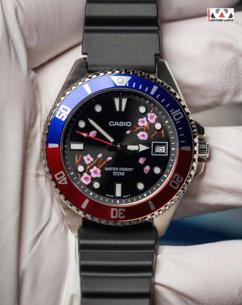 May include: A Casio watch with a black face, pink floral details, and a date display. It has a stainless steel case, a black rubber strap, and a rotating bezel with blue and red sections. The watch face displays "CASIO" and "WATER RESIST 50M".