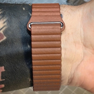 May include: A brown leather watch band with a ribbed design and a silver-colored clasp. The band is shown on a wrist, showcasing its texture and color. The watch band is a stylish accessory.
