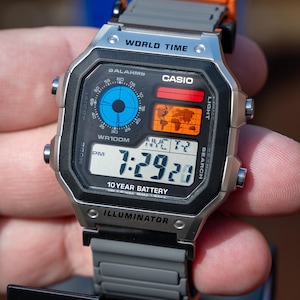 May include: A digital Casio watch with a silver case and grey strap. The watch face shows the time, world time, and other functions. The watch has an orange clasp and is labelled "WORLD TIME" and "ILLUMINATOR".