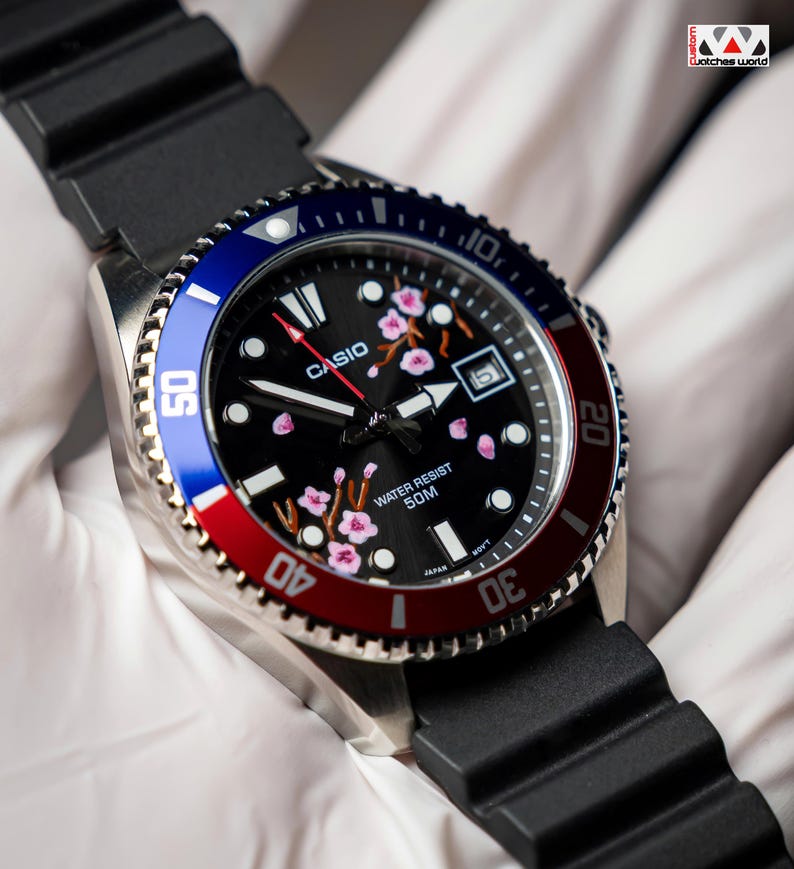 May include: A Casio watch with a black face featuring pink floral accents. The watch has a blue and red bezel, a black rubber strap, and the text "CASIO" and "WATER RESIST 50M".