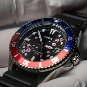 May include: A Casio watch with a black face featuring pink cherry blossom designs. The watch has a red and blue rotating bezel and a black rubber strap. The watch face displays the Casio logo and the words "Water Resist 50M".
