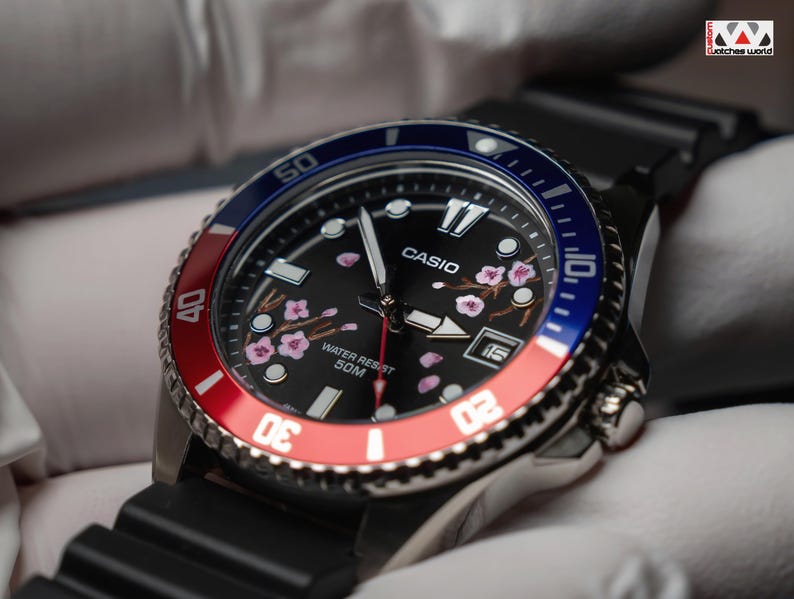 May include: A Casio watch with a black face featuring pink cherry blossom designs. The watch has a red and blue rotating bezel and a black rubber strap. The watch face displays the Casio logo and the words "Water Resist 50M".