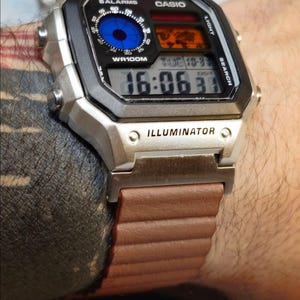 May include: A silver Casio digital watch with a black face and brown band. The watch face displays the time, date, and other functions. The watch has the word "ILLUMINATOR" printed on the face.