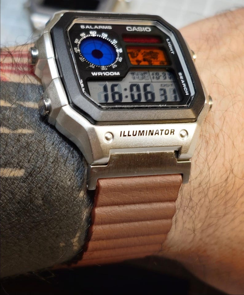 May include: A silver Casio digital watch with a black face and brown band. The watch face displays the time, date, and other functions. The watch has the word "ILLUMINATOR" printed on the face.