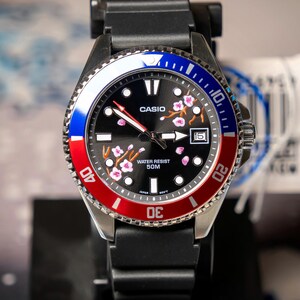 May include: A black Casio watch with a red, white, and blue bezel. The watch face is black with pink floral accents and the Casio logo. The watch has a black band and a date display at the 3 o'clock position. The watch is water resistant to 50M.