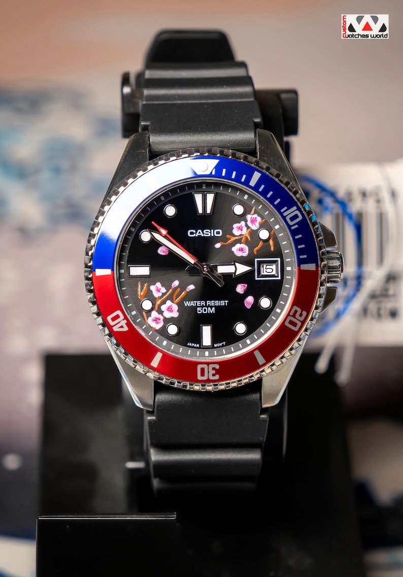 May include: A black Casio watch with a red, white, and blue bezel. The watch face is black with pink floral accents and the Casio logo. The watch has a black band and a date display at the 3 o'clock position. The watch is water resistant to 50M.