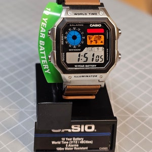 May include: A Casio digital watch with a silver case, brown leather strap, and a green tag that reads "10 YEAR BATTERY." The watch face displays the time, date, and world map. The watch is on a black display stand.