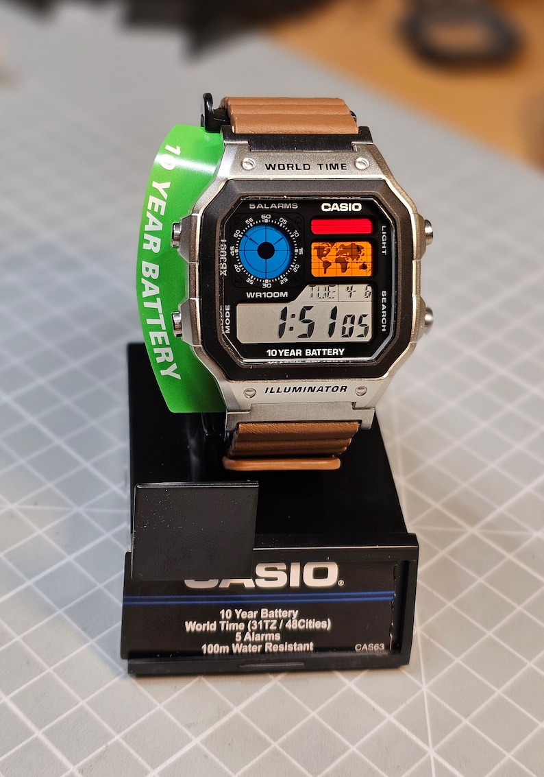 May include: A Casio digital watch with a silver case, brown leather strap, and a green tag that reads "10 YEAR BATTERY." The watch face displays the time, date, and world map. The watch is on a black display stand.