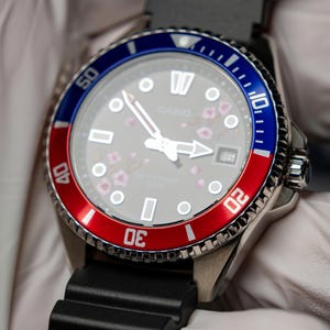 May include: A stainless steel wristwatch with a black rubber strap. The watch face is gray with white markers and hands, and a date window at the 3 o'clock position. The bezel is red and blue, and the watch has a floral design.