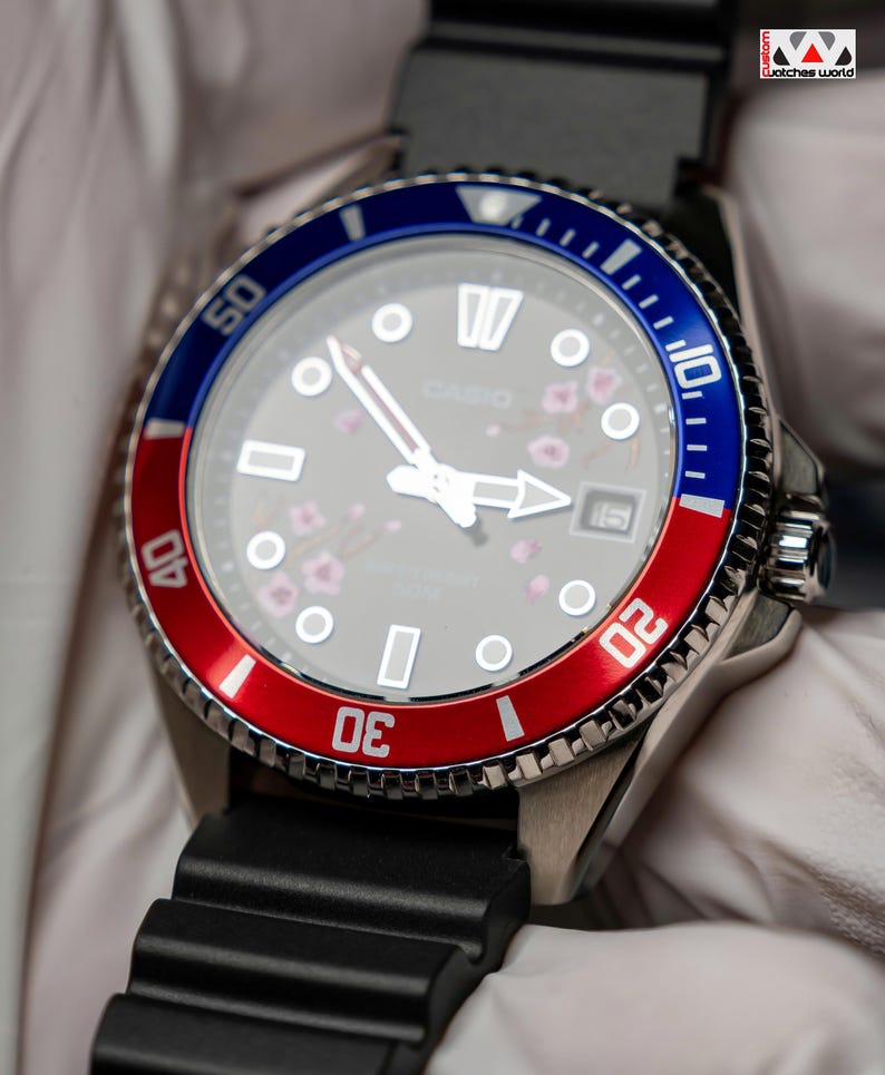 May include: A stainless steel wristwatch with a black rubber strap. The watch face is gray with white markers and hands, and a date window at the 3 o'clock position. The bezel is red and blue, and the watch has a floral design.
