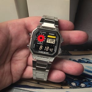 May include: A silver Casio watch with a black face and a red alarm indicator. The watch has a digital display that shows the time, date, and other features. The watch also has a world time function and a 10-year battery.