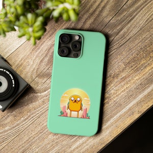 Puede incluir: Funda de teléfono verde menta con un personaje de dibujos animados en un paisaje desértico. El personaje amarillo tiene una expresión triste, frente a una puesta de sol. La funda tiene un recorte para la cámara y está sobre una superficie de madera.