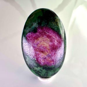 Puede incluir: Una piedra preciosa pulida de forma ovalada con un centro rosa magenta vibrante rodeado de inclusiones minerales de color verde bosque profundo y negro. La superficie es lisa y reflectante, mostrando un contraste natural entre el verde oscuro y el rosa.