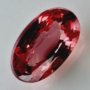 May include: An oval-cut gemstone with a vibrant reddish-pink hue and a faceted surface that reflects light. The stone displays deep crimson tones and lighter pink highlights throughout its transparent structure, showcasing a clear, polished finish.
