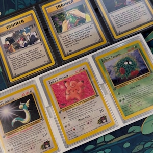 May include: A collection of vintage Pokémon trading cards, including Trainer and Pokémon cards. The cards are in protective sleeves and feature characters like Erika's Clefairy, Dratini, and Tangela. The cards are displayed on a patterned surface.