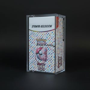 May include: A clear acrylic display case containing a "Booster Bundle" of "Pokemon Trading Card Game" cards. The cards feature a pink Mew character and the text "151". The case is on a black surface.