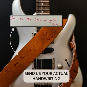 Pu&ograve; includere: Una chitarra elettrica argentata con un cinturino per chitarra in pelle marrone. Il cinturino presenta un testo scritto a mano: "You are the love of my life" con un cuore e il nome "Sarah". L'immagine include anche il testo "SEND US YOUR ACTUAL HANDWRITING."