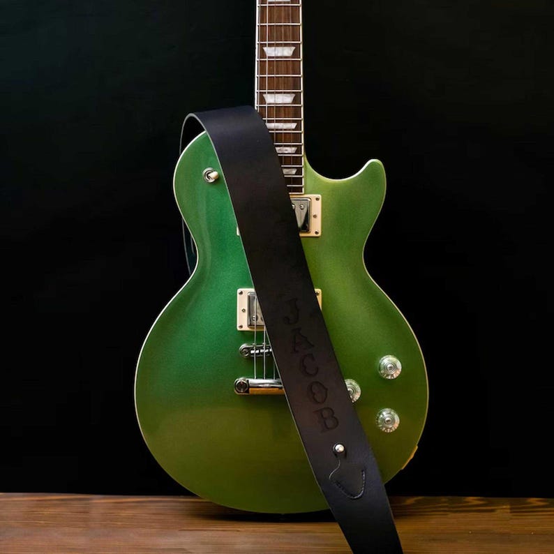 Pu&ograve; includere: Una chitarra elettrica verde con un cinturino in pelle nera con il nome "JACOB". La chitarra ha hardware argentato ed &egrave; su uno sfondo scuro. La chitarra misura circa 99 cm di lunghezza.