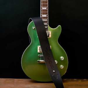 May include: A green electric guitar with a black leather strap. The strap is draped over the guitar and features the name "JACOB" embossed in capital letters. The guitar has silver hardware and is set against a dark background.