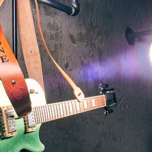 May include: A green electric guitar with a brown leather strap, the strap is embossed with the word "ZONE". The guitar is set against a dark, textured wall with a bright spotlight.