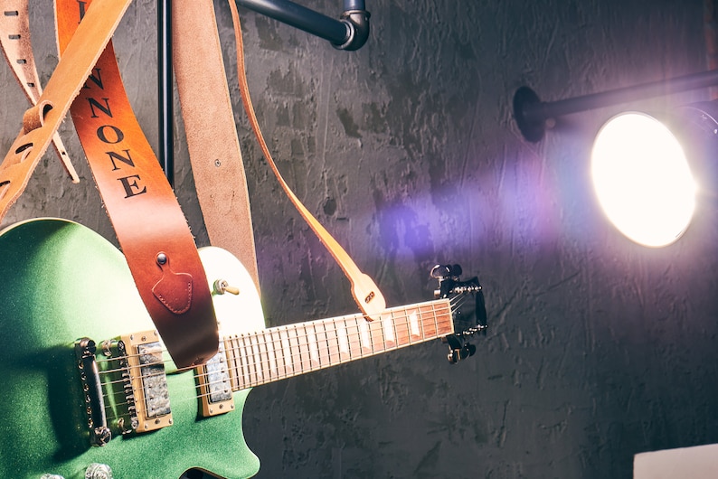 May include: A green electric guitar with a brown leather strap, the strap is embossed with the word "ZONE". The guitar is set against a dark, textured wall with a bright spotlight.