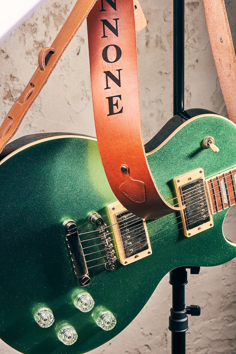 Pu&ograve; includere: Una chitarra elettrica verde con finitura scintillante, abbinata a un cinturino per chitarra in pelle marrone. La parola "ZONE" &egrave; stampata in nero sul cinturino. La chitarra ha pickup color crema e hardware cromato.