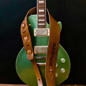 May include: A green electric guitar with a brown leather guitar strap. The strap is embossed with the name "PETER". The guitar has silver knobs and pickups. The background is black.