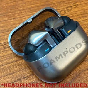 May include: A silver and black wireless headphone charging case with the word "FOAMPODS" printed on the side.