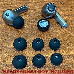 May include: A set of seven black earbud tips for headphones. The tips are of varying sizes and are arranged in a row on a wooden surface.