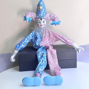 May include: A porcelain doll dressed as a jester with a blue and pink star patterned costume. The doll is wearing a blue and white hat with a bell on top. The doll is sitting on a black box.