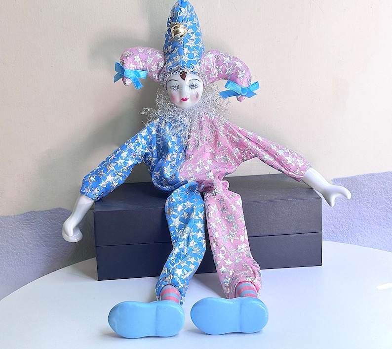 May include: A porcelain doll dressed as a jester with a blue and pink star patterned costume. The doll is wearing a blue and white hat with a bell on top. The doll is sitting on a black box.