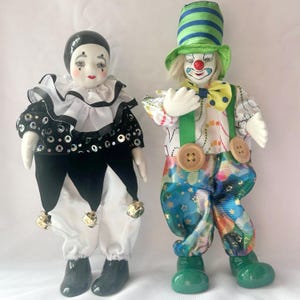 May include: Two decorative clown dolls. One doll wears a black and white harlequin outfit with bells, the other wears a colourful outfit with a green hat and braces. Both dolls have painted faces.