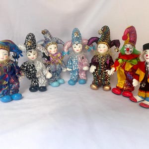 May include: A collection of eight porcelain clown dolls, each with a unique colorful costume and jester hat. The dolls are arranged in a row on a white surface.