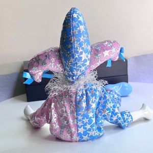 May include: A blue and pink jester doll with a star pattern and a white lace collar. The doll is sitting on a white surface.