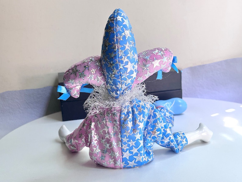 May include: A blue and pink jester doll with a star pattern and a white lace collar. The doll is sitting on a white surface.