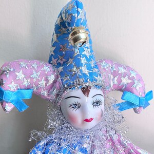 May include: A porcelain jester doll with a blue and pink star patterned hat and a silver bell. The doll has a white face with painted features and a red mouth.
