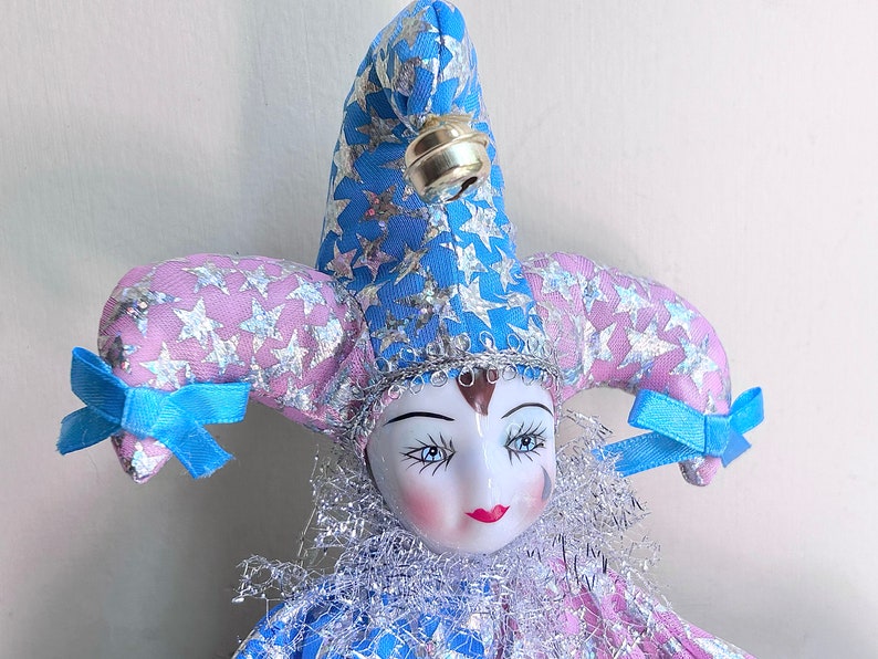 May include: A porcelain jester doll with a blue and pink star patterned hat and a silver bell. The doll has a white face with painted features and a red mouth.