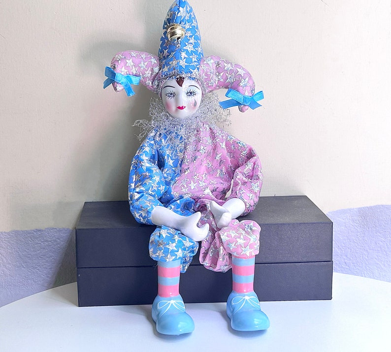 May include: A porcelain jester doll with a blue and pink star patterned costume. The doll has a blue and white hat with a bell on top. The doll is sitting on a black box.
