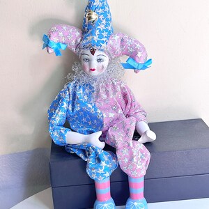 May include: A porcelain jester doll with a blue and pink star patterned costume, a blue and white striped hat with a bell, and blue shoes with white bows.